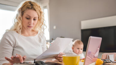 Women with blond hair working with papers and a laptop at home with baby nearby.