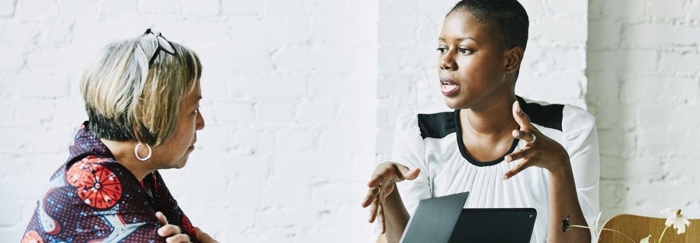 A white woman and a Black woman are sitting at a table talking in front of a laptop computer