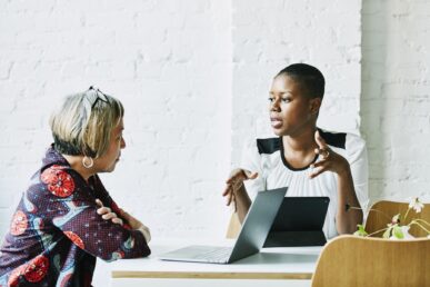 A white woman and a Black woman are sitting at a table talking in front of a laptop computer