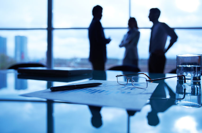 Close-up of business document, pen, glass of water and eyeglasses at workplace on background of office workers interacting